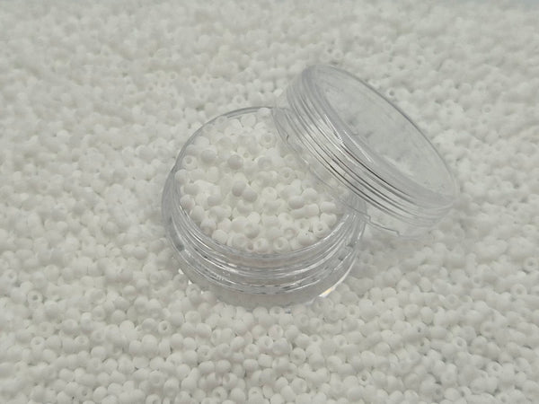 Små, runde, matte hvide perler i en gennemsigtig plastikbeholder, som står på en masse perler af samme art. 