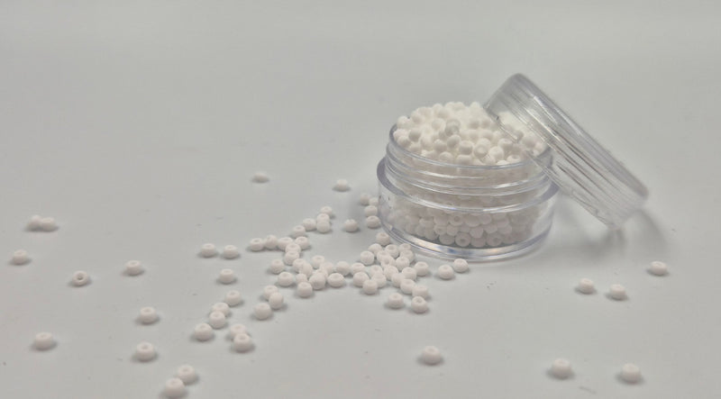 Små, runde, matte hvide perler i en gennemsigtig plastikbeholder, hvor de falder lidt ud af, og ligger på en grå baggrund. 