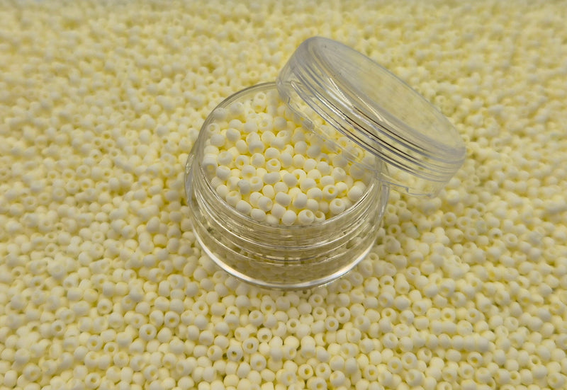 Små, runde, matte lysegule perler i en gennemsigtig plastikbeholder, som står på en masse perler af samme art. 
