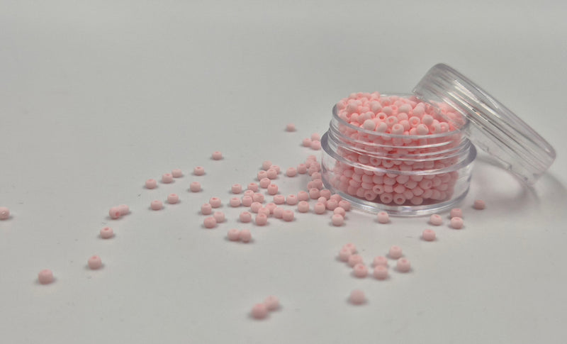 Små, runde, matte lyserøde perler i en gennemsigtig plastikbeholder, hvor de falder lidt ud af, og ligger på en grå baggrund. 