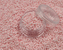 Små, runde, matte lyserøde perler i en gennemsigtig plastikbeholder, som står på en masse perler af samme art. 