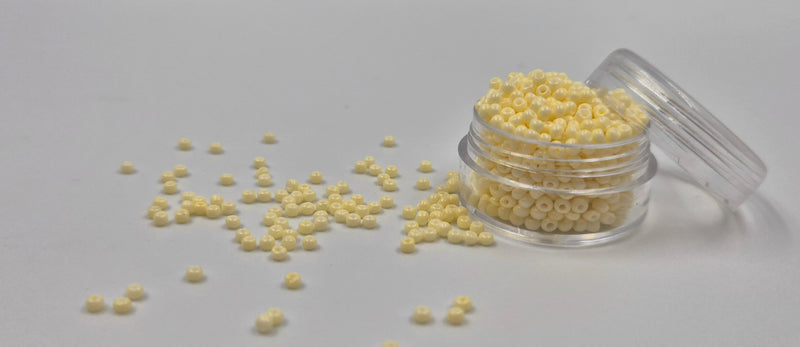 Små, runde, elfenbens-farvede perler i en gennemsigtig plastikbeholder, hvor de falder lidt ud af, og ligger på en grå baggrund. 