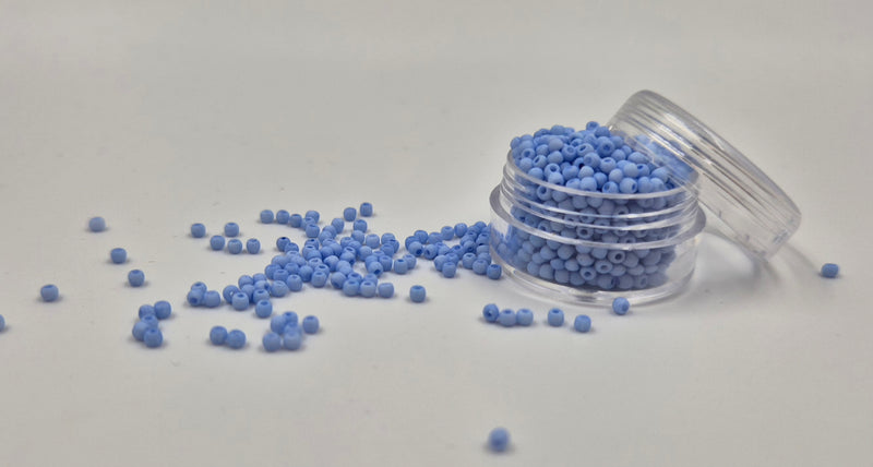 Små, runde, matte pastelblå perler i en gennemsigtig plastikbeholder, hvor de falder lidt ud af, og ligger på en grå baggrund. 
