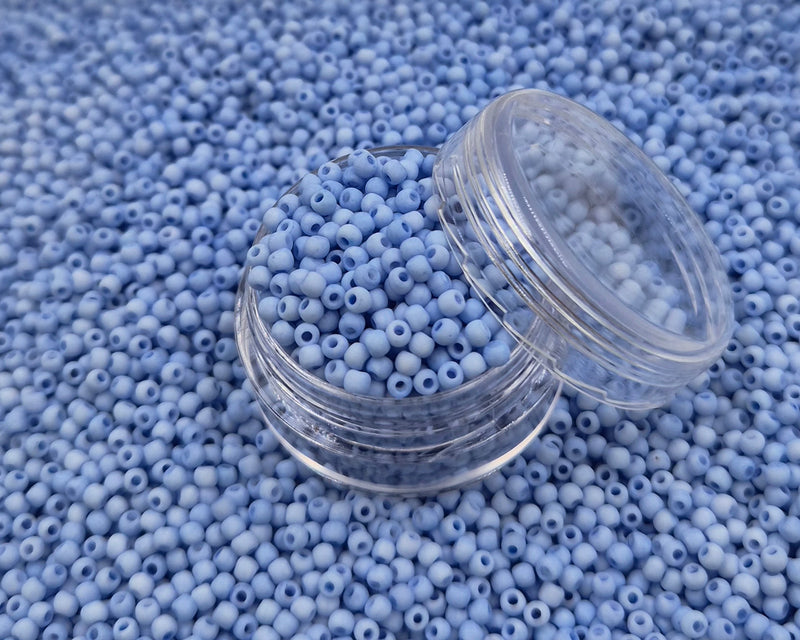 Små, runde, matte pastelblå perler i en gennemsigtig plastikbeholder, hvor de falder lidt ud af, og ligger på en grå baggrund. 