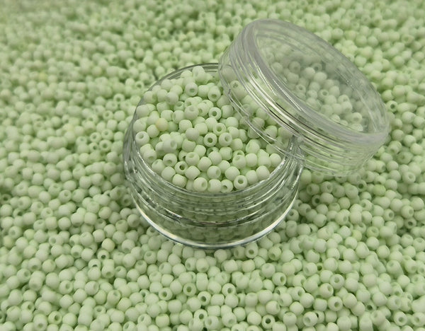 Små, runde, matte pastelgrønne perler i en gennemsigtig plastikbeholder, hvor de falder lidt ud af, og ligger på en grå baggrund. 