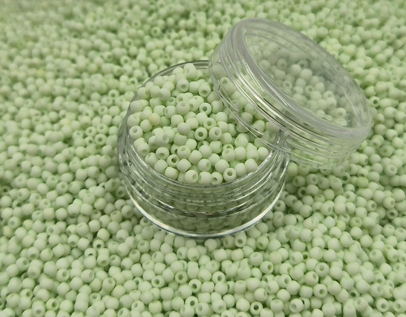 Små, runde, matte pastelgrønne perler i en gennemsigtig plastikbeholder, hvor de falder lidt ud af, og ligger på en grå baggrund. 