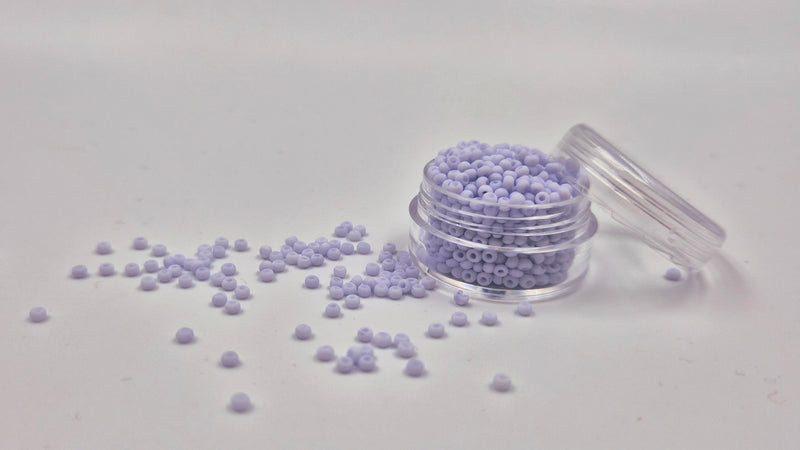 Små, runde, matte pastellavendel perler i en gennemsigtig plastikbeholder, hvor de falder lidt ud af, og ligger på en grå baggrund. 