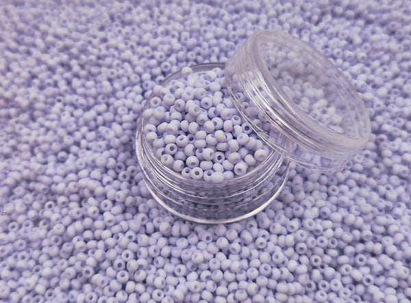 Små, runde, matte pastellavendel perler i en gennemsigtig plastikbeholder, hvor de falder lidt ud af, og ligger på en grå baggrund. 