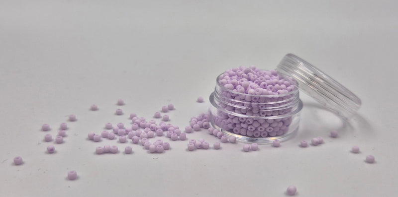 Små, runde, matte pastel-lilla perler i en gennemsigtig plastikbeholder, hvor de falder lidt ud af, og ligger på en grå baggrund. 