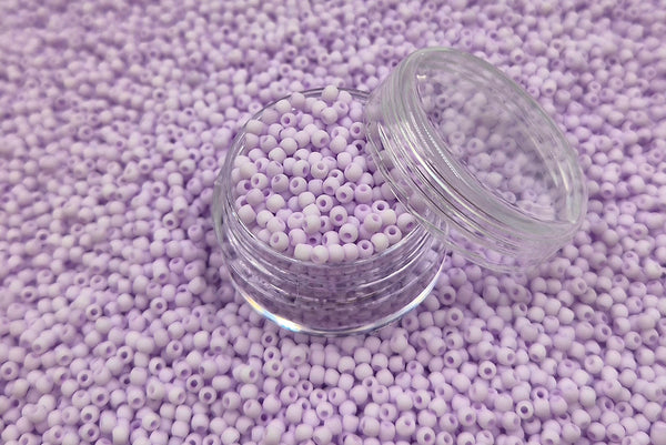 Små, runde, matte pastel-lilla perler i en gennemsigtig plastikbeholder, hvor de falder lidt ud af, og ligger på en grå baggrund. 