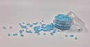 Små, runde, matte pastelturkis perler i en gennemsigtig plastikbeholder, hvor de falder lidt ud af, og ligger på en grå baggrund. 