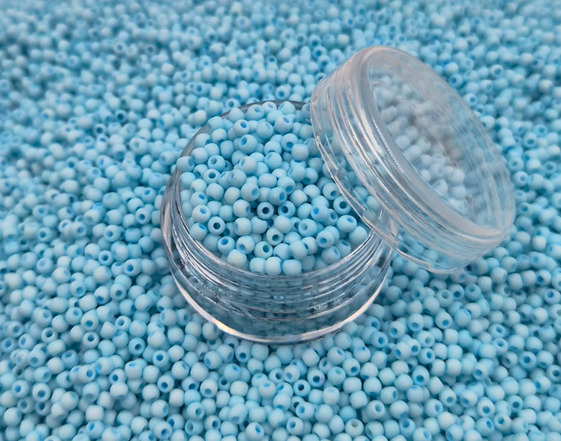 Små, runde, matte pastelturkis perler i en gennemsigtig plastikbeholder, hvor de falder lidt ud af, og ligger på en grå baggrund. 