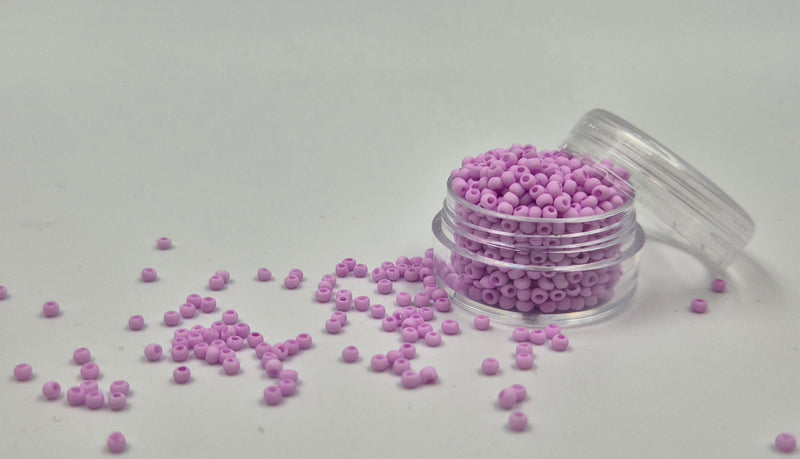 Små, runde, matte pastel-rosa perler i en gennemsigtig plastikbeholder, hvor de falder lidt ud af, og ligger på en grå baggrund. 