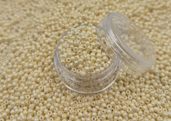 Små, runde, perlemor elfenbens-farvede perler i en gennemsigtig plastikbeholder, som står på en masse perler af samme art. 