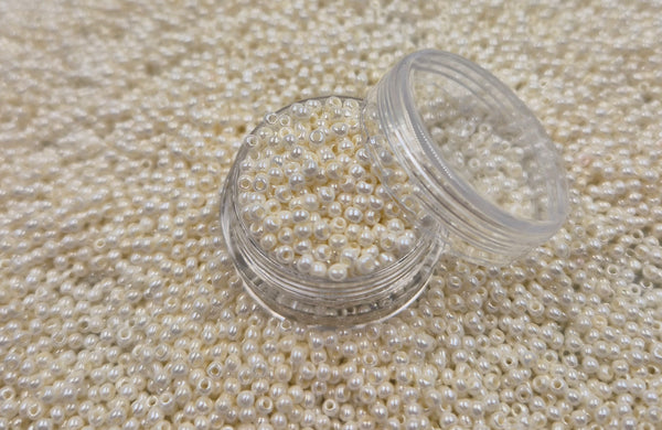 Små, runde, hvide perlemor perler i en gennemsigtig plastikbeholder, som står på en masse perler af samme art. 