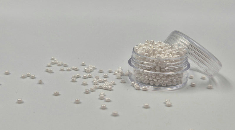 Små, runde, skinnende-hvide perler i en gennemsigtig plastikbeholder, hvor de falder lidt ud af, og ligger på en grå baggrund. 