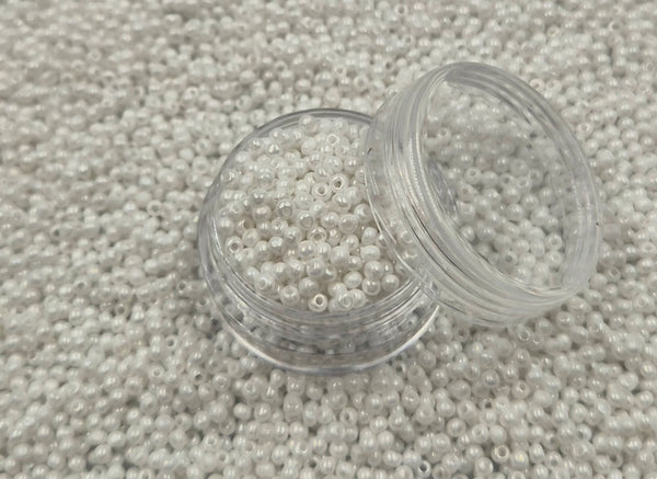 Små, runde, skinnende-hvide perler i en gennemsigtig plastikbeholder, hvor de falder lidt ud af, og ligger på en grå baggrund. 