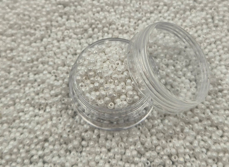 Små, runde, skinnende-hvide perler i en gennemsigtig plastikbeholder, hvor de falder lidt ud af, og ligger på en grå baggrund. 