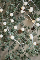 Et stemningsbillede med en lyskæde, gran med grankogler i plastik, som hænger op ad en væg. 