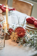 Et stemningsbillede af en bordopsætning til jul, med røde kugler, steatinlys, gran guirlande samt tallerkner med røde stofservietter på, og glas foran. 